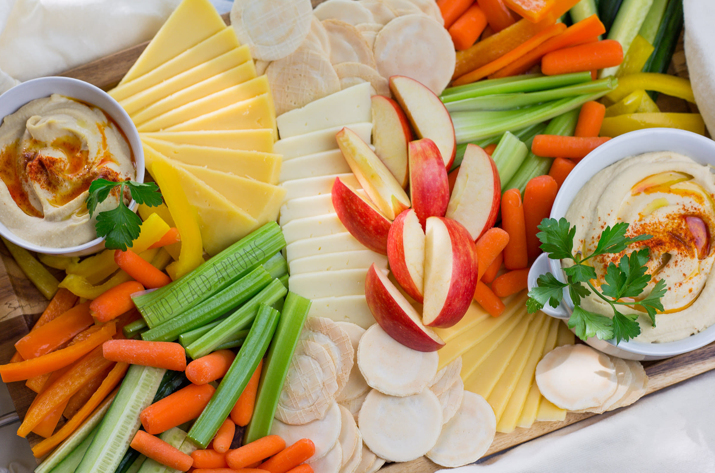 Veggie board filled with two bowls of hummus, cheeses, carrots, celery, cucumber, bell peppers, and rice crackers.