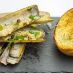 Cooked razor clams stacked on black graphite slate with toasted bread on right.