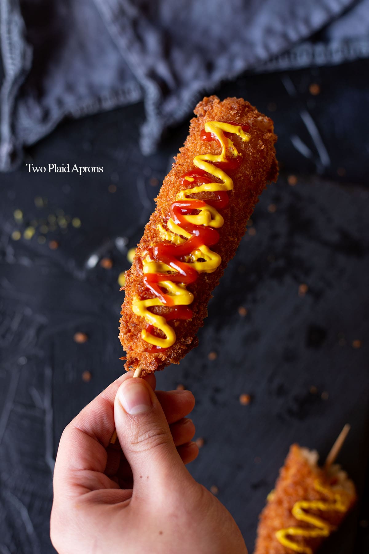 Holding a Korean corn dog drizzled with ketchup and yellow mustard.