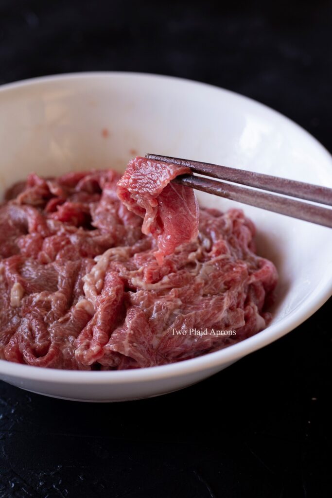 A bowl of marinating sliced beef with a pair of chopsticks holding up a slice for reference.